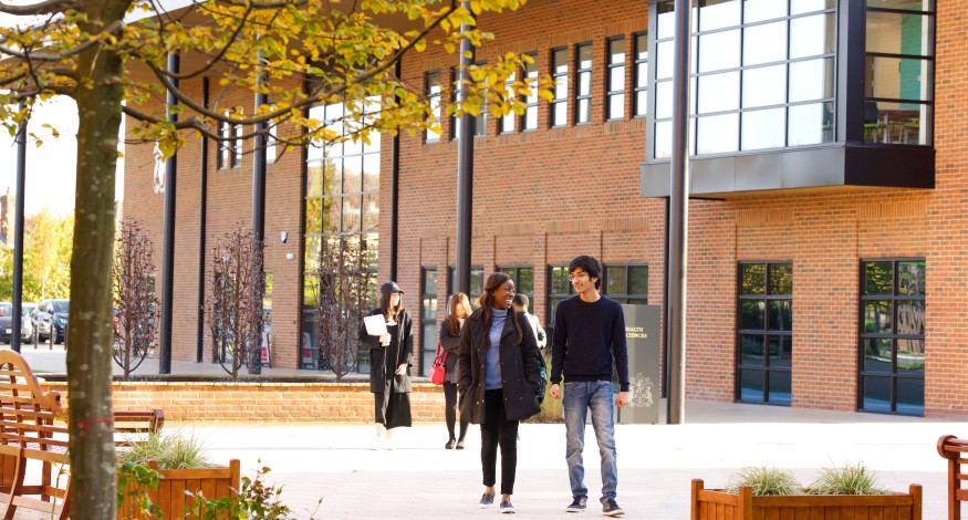 A male and female student walking under a tree in front of a building.